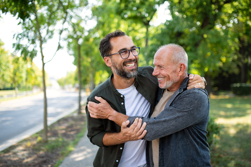 Two men with glasses are hugging each other outdoors in front of a tree, both smiling and wearing casual clothing.