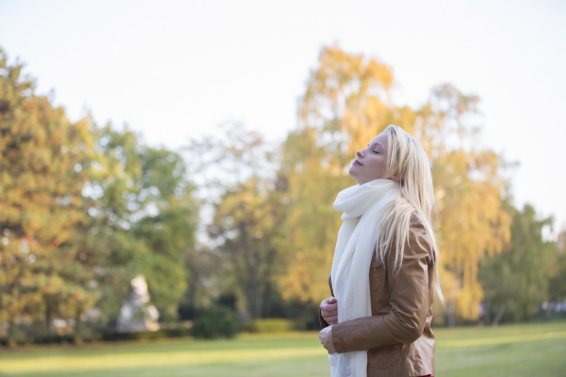 A woman wearing a brown jacket and a white scarf stands in a park surrounded by trees and grass under a clear sky, with autumn leaves scattered around.
