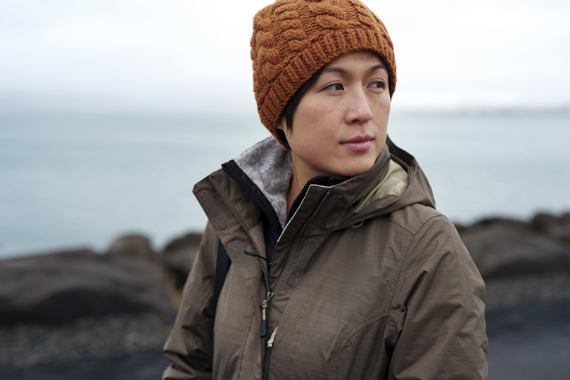A woman wearing a hat and a jacket stands outdoors, with winter clothing including a scarf. She is at the beach.