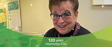 Kathy smiling sitting at her desk with a visual overlay "120 years improving lives"