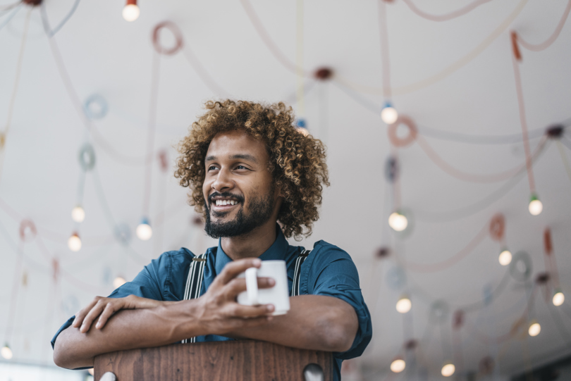 A man with curly hair and a beard smiles warmly while holding a white mug. Soft, colorful hanging lights create a relaxed, cheerful atmosphere.
