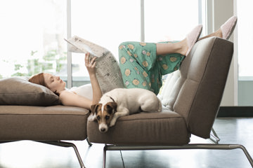 Woman relaxes on sofa with newspaper and dog, preserving her energy in case of migraine