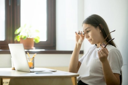 Woman working at laptop, struggling with migraine-triggering eyestrain when not wearing her glasses.