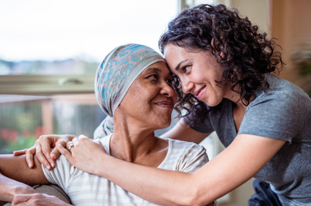 A woman in a headscarf and striped shirt is lovingly embraced by a younger woman with curly hair. They exchange warm smiles near a sunlit window.