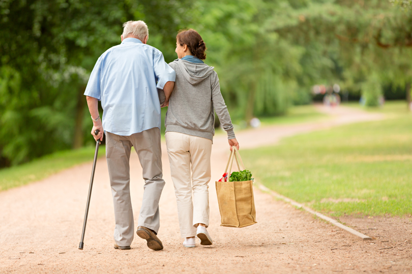 Elderly man with cane and younger woman carrying groceries walk arm-in-arm on a tree-lined park path. Tone conveys support and companionship.