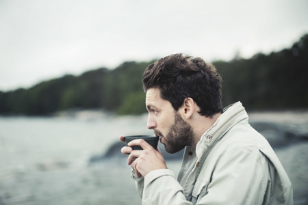 A man in a light jacket sips from a mug by a calm lakeside with a forested background. The mood is serene and contemplative.