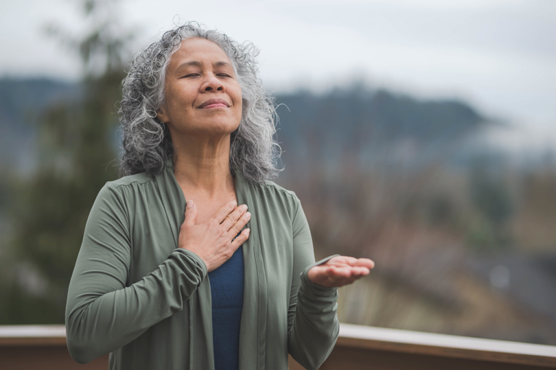 An older woman with gray curly hair stands outdoors, eyes closed, with one hand on her chest and the other open in front of her, appearing calm and relaxed.