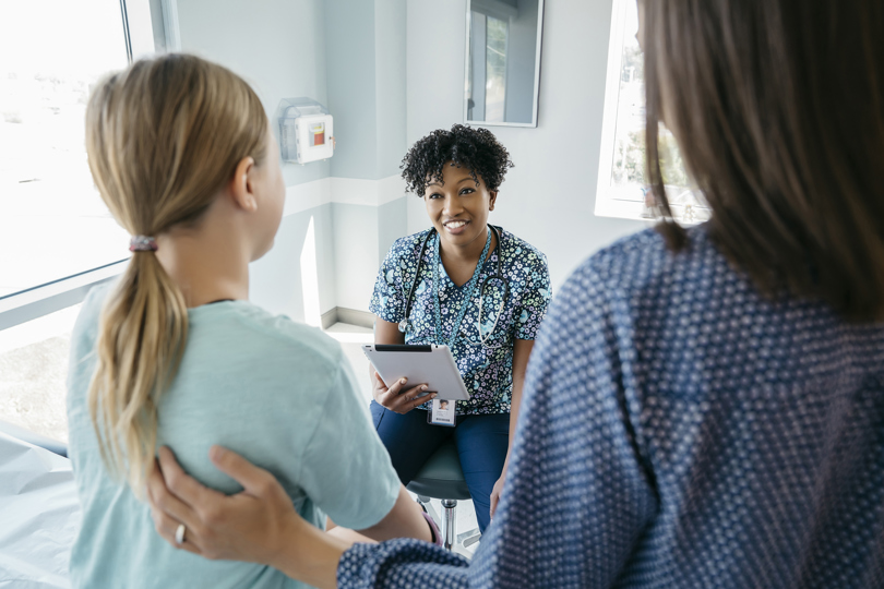 A smiling nurse in scrubs with a stethoscope talks to a young girl and an adult in a clinic. The scene conveys warmth and care.