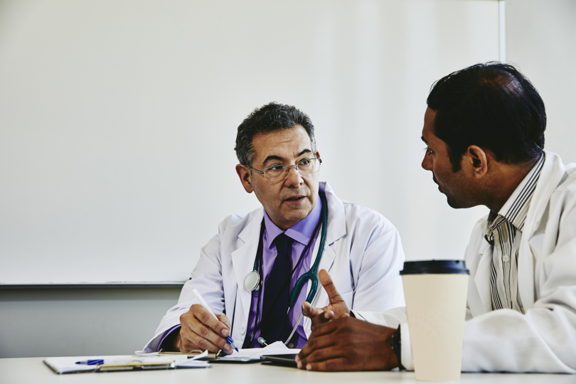Two doctors in white coats have a focused discussion at a table. One holds a pen, while the other gestures. A coffee cup sits nearby.