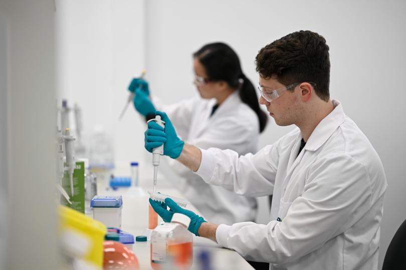 Two scientists in lab coats and safety glasses use pipettes to transfer liquids into test tubes in a laboratory setting.