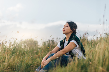 A woman sits peacefully in a grassy field at sunset, looking upwards with a serene expression. The sky is partly cloudy, creating a tranquil atmosphere.